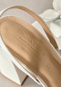 Bijela sandala na petu s teksturiranom kožnom trakom. Unutrašnjost sadrži utisnuti brend: "SIGNATURE MADE IN BRAZIL NEXT." Veličina: 6.5/40.