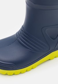 Botte en caoutchouc bleu marine avec une texture striée autour de la cheville et une semelle robuste jaune vif pour une utilisation en extérieur.