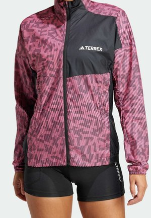 Frau trägt eine rosa-schwarz gemusterte Terrex-Jacke mit Frontreißverschluss und schwarze Terrex-Radshorts.
