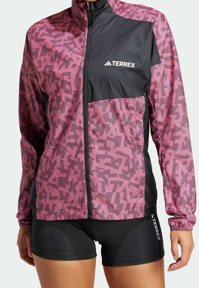 Femme portant une veste Terrex rose et noire à motifs avec une fermeture éclair à l'avant et un short de cyclisme Terrex noir.