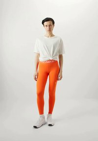 Oransje leggings med en hvit Nike-logo på linningen, kombinert med en lys grå knyttet t-skjorte og hvite treningssko.