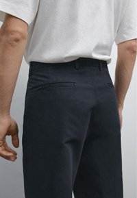 Pantaloni in cotone blu scuro con una vestibilità su misura, dotati di due tasche posteriori e una texture liscia. Abbinati a una semplice maglietta bianca.