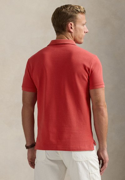 Bărbat blond văzut din spate, purtând un tricou polo roșu cu mâneci scurte și pantaloni albi, stând în fața unui fundal neutru.