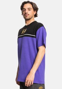 Fialové a černé tričko s krátkým rukávem a kulatým výstřihem, s bílým pruhem a žlutým logem na hrudi. Texturovaná tkanina.