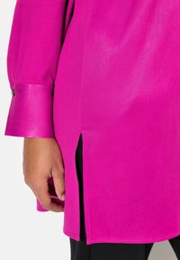 Fuchsia Bluse mit seidiger Textur, verfügt über einen seitlichen Schlitz und einen knöpfbaren Ärmelabschluss. Hand in der Nähe des Saums positioniert.