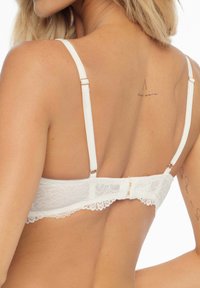 Weißer Spitzen-BH mit verstellbaren Trägern und Hakenverschluss, der ein zartes Blumenmuster entlang des Bandes und eine glatte Textur aufweist.