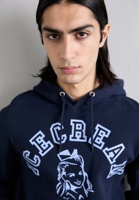 Navy blue hoodie featuring a graphic design of a woman’s face and bold white lettering. Made of soft fabric with a drawstring hood.