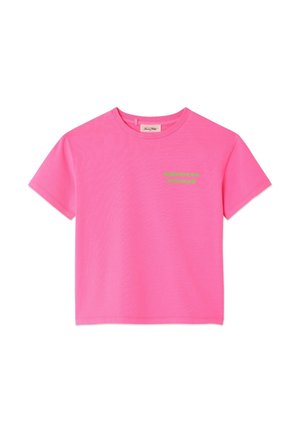 Enfant aux cheveux bouclés portant un t-shirt rose vif avec l'inscription "AMERICAN VINTAGE" dans le dos et un pantalon gris clair, debout à l'intérieur.