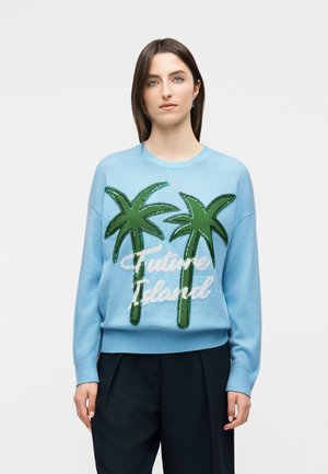 Femme aux cheveux bruns raides portant un pull bleu clair avec deux palmiers verts et le texte "Future Island", associé à un pantalon noir.