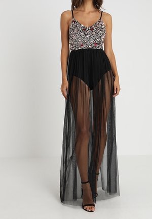 Vrouw in een mouwloze jurk met een bloemenborduurwerk op het lijfje en een lange doorzichtige zwarte rok over zwarte shorts, staand op een witte vloer.