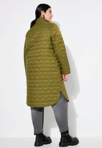 Cappotto trapuntato verde oliva con motivo a cuori, maniche lunghe e spacchi laterali. Abbinato a jeans grigi e stivaletti neri.