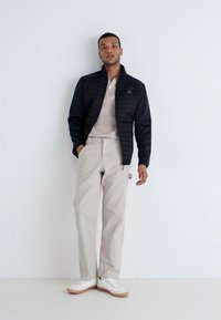 Sort polstret jakke, beige rippede sweater, beige bukser og hvide sneakers med gummisåler. Model står foran en ensfarvet baggrund.