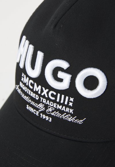 Casquette noire en tissu ornée du texte brodé blanc "HUGO", avec des éléments décoratifs incluant l'année "1993" et des détails de marque.