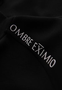 Tela negra con texto blanco que dice "OMBRE EXIMIO." Textura suave con un diseño moderno y minimalista.