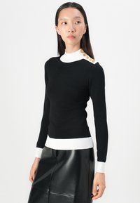 Zwarte trui met een hoge witte kraag, voorzien van gouden knopen op één schouder en witte geribde manchetten en onderkant. Geweven textiel.