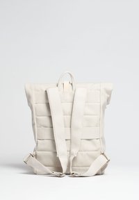 Ljusbeige ryggsäck med quiltad textur, justerbara tygband, bärhandtag och en minimalistisk design. Detaljerna inkluderar metallklämmor.