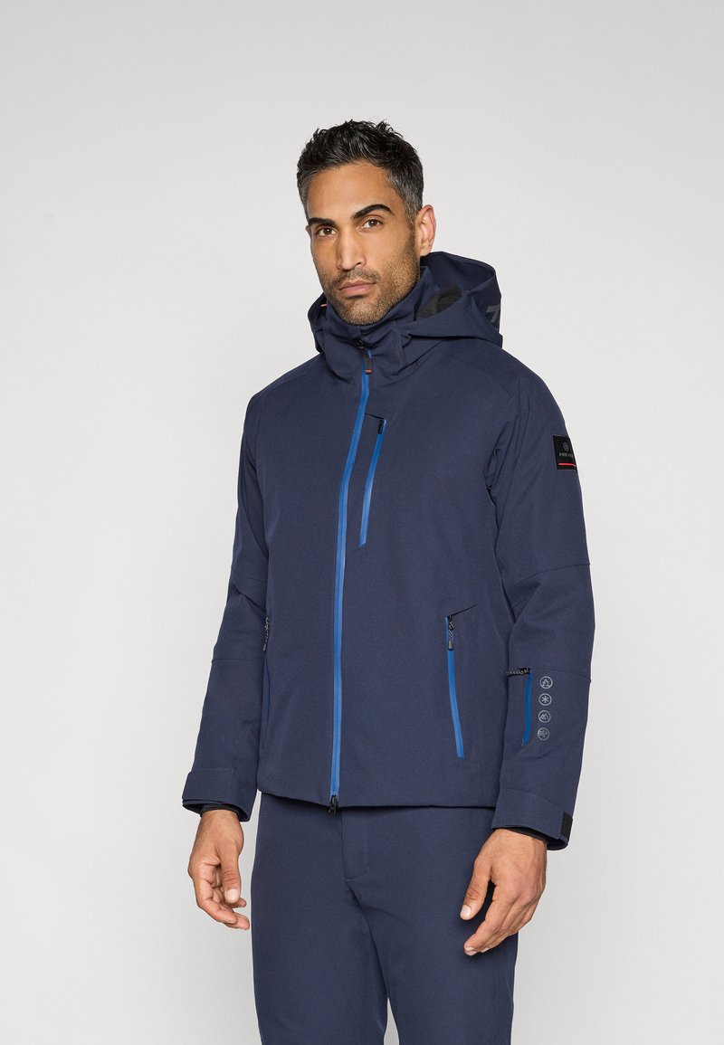 Tamsiai mėlyna neperšlampama striukė su gobtuvu, turi mėlynas užtrauktukus, kišenes užtrauktuku ir logotipo pleistrą ant rankovės. Lygi tekstūra.