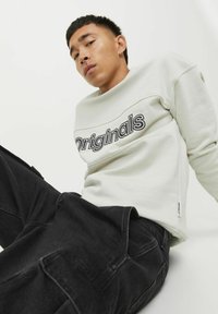 Sudadera gris claro con "Originals" impreso en negrita negra, combinada con pantalones cargo negros; textura suave y corte holgado.