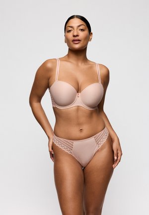 Vrouw met een medium huidskleur die een naaktkleurige beha met dubbele bandjes en bijpassende slip met kanten randjes draagt, staand tegen een effen lichtgrijze achtergrond.