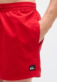 Ερυθρά μαγιό σορτς με ελαστική μέση, πλαϊνές τσέπες, λεία υφή και μαύρο λογότυπο στο τελείωμα.