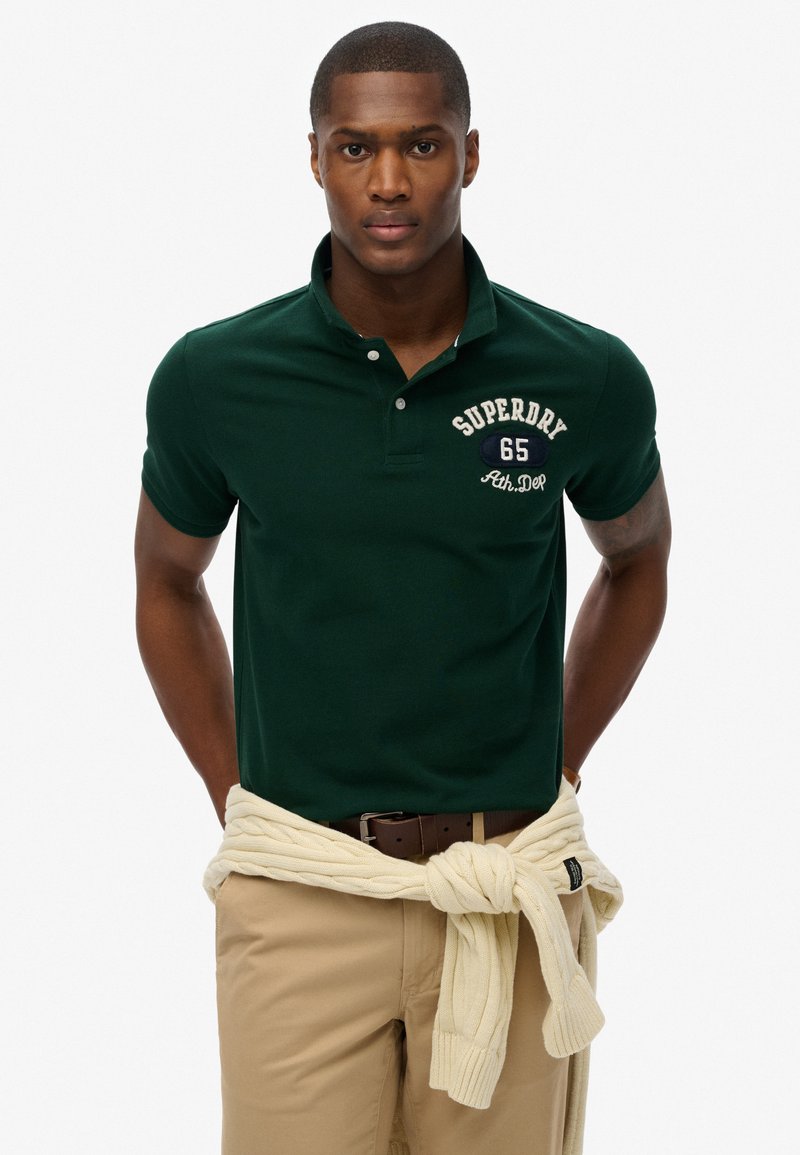 Polo vert avec un col, ornée d'une broderie blanche "SUPERDRY 65". Associé à un pull crème noué à la taille et un pantalon beige.