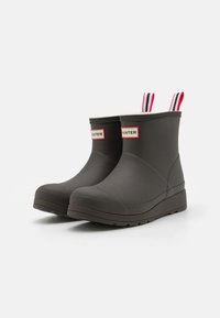 Bottes de cheville en caoutchouc noir avec semelles plates, forme courbée et languettes de tirage rayées rouges et blanches à l'arrière. Présente le logo de la marque sur les côtés.