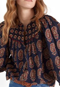 Blusa blu navy con stampa paisley in rosso e crema. Caratterizzata da un collo arricciato, maniche voluminose e chiusura con bottoni. Tessuto testurizzato.