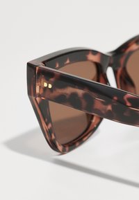 Óculos de sol padrão tartaruga com lente degradê castanho escuro, design de armação angular e detalhes em dourado nas hastes.