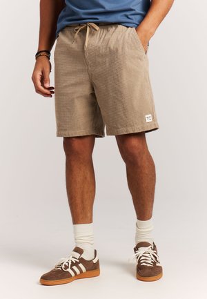 Man wearing beige checkered shorts with drawstring, blue shirt, white socks, and brown sneakers standing against a plain background.
