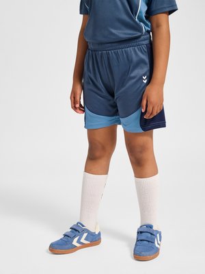 Kind trägt blaue Sportshorts, passende Sneakers mit weißen Chevron-Logos, weiße knielange Socken, steht vor einem grauen Hintergrund.