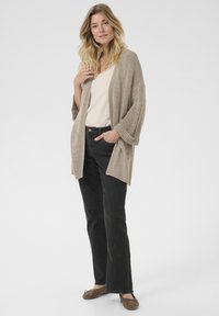 Beige gestrickte Strickjacke mit lockeren Ärmeln über einem hellen Oberteil, kombiniert mit dunklen Jeans und braunen Mokassins. Neutrale Farbpalette.
