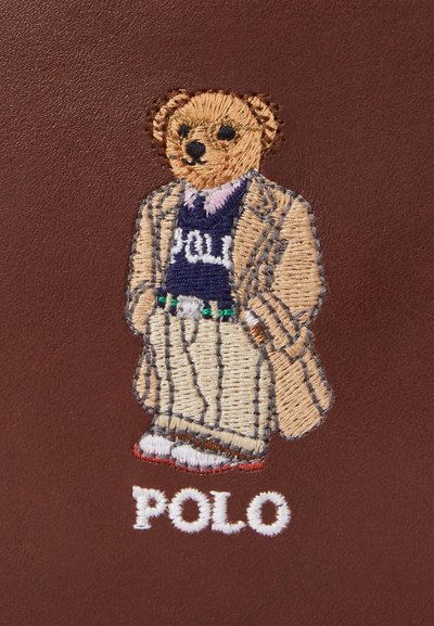 Hímzett medve, aki bézs csíkos öltönyöt és sötétkék pulóvert visel, rajta a "Polo" felirattal, barna háttér előtt.