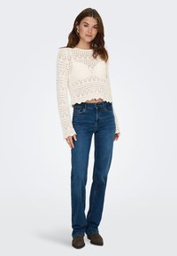 Haut court en crochet crème avec manches longues et ourlet festonné. Associé à un jean slim en denim bleu foncé et des bottines marron.