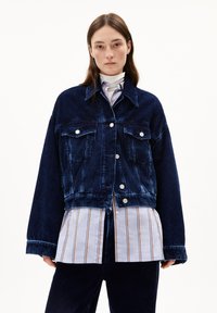 Donna che indossa una giacca di velluto a coste blu scuro oversize sopra una maglietta a righe e un dolcevita bianco, in piedi contro uno sfondo bianco semplice.