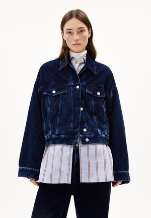 Vrouw in een oversized donkerblauwe corduroy jas over een gestreept shirt en witte coltrui, staand tegen een effen witte achtergrond.
