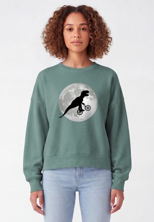 Femme aux cheveux bouclés portant un sweat vert avec une silhouette noire d'un dinosaure à vélo devant une pleine lune.