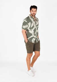 Camisa de manga corta en verde con estampado de hojas blancas, combinada con pantalones cortos cargo en verde oliva y zapatillas blancas. Ajuste casual y tejido ligero.