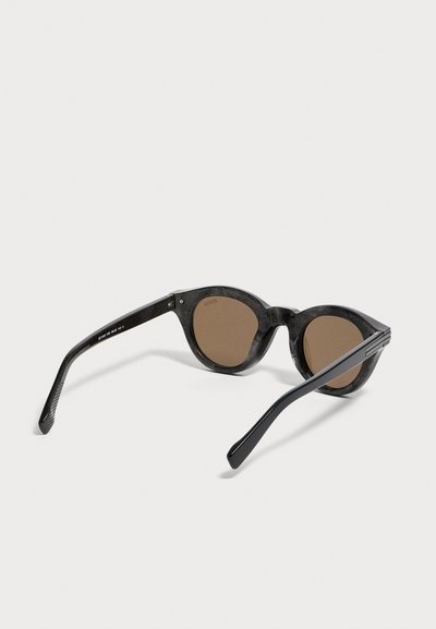 Lunettes de soleil noires à monture ronde avec verres marron foncé, inclinées pour montrer l'avant et les branches latérales sur fond blanc.