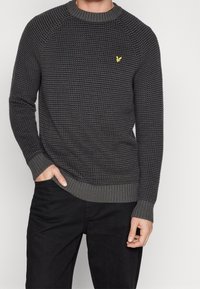 Dark gray knitted sweater with a textured pattern, ribbed collar and cuffs, and a small yellow logo on the chest.
