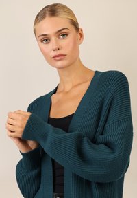Cardigan a coste blu verde con apertura frontale, spalle abbassate e maniche lunghe. Indossato sopra un top nero aderente, che mostra un motivo testurizzato.