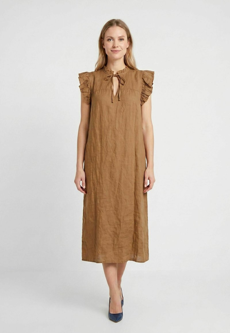 Femme portant une robe marron mi-longue avec des manches volantées et un lien au niveau du col, assortie à des escarpins bleu foncé, debout sur fond blanc.