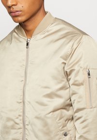Giacca bomber beige realizzata in un tessuto liscio e lucido, con colletto a costine, tasche laterali e tasca con zip sulla manica impreziosita da dettagli in metallo argentato.