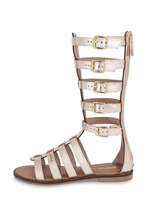 Sandalias gladiador con tiras en cuero metálico beige. Presentan múltiples hebillas, punta abierta y cierre con cremallera en la parte trasera.