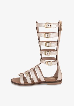 Sandalias gladiador con tiras en cuero metálico beige. Presentan múltiples hebillas, punta abierta y cierre con cremallera en la parte trasera.