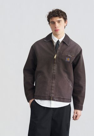 Joven con cabello rizado que lleva una chaqueta marrón oscuro con cremallera sobre una camisa blanca y corbata negra, combinada con pantalones cargo negros, frente a un fondo liso.