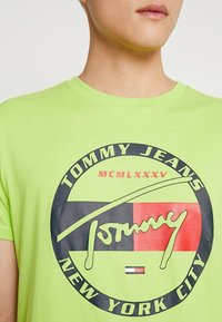 Jasnozielona bawełniana koszulka z okrągłym czarnym i czerwonym logo. Zawiera tekst "TOMMY JEANS" oraz "NEW YORK CITY" w pogrubionej czcionce.