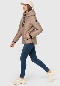 Light brown padded jacket with a high collar, black zipper and accents, worn with blue jeans and light-colored sneakers, with a beige cap.