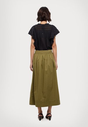 Frau mit kurzem dunklem Haar, die eine schwarze Spitzenbluse, einen olivgrünen Midi-Rock und schwarze Sandalen mit Absatz trägt, steht mit dem Rücken zur Kamera vor weißem Hintergrund.