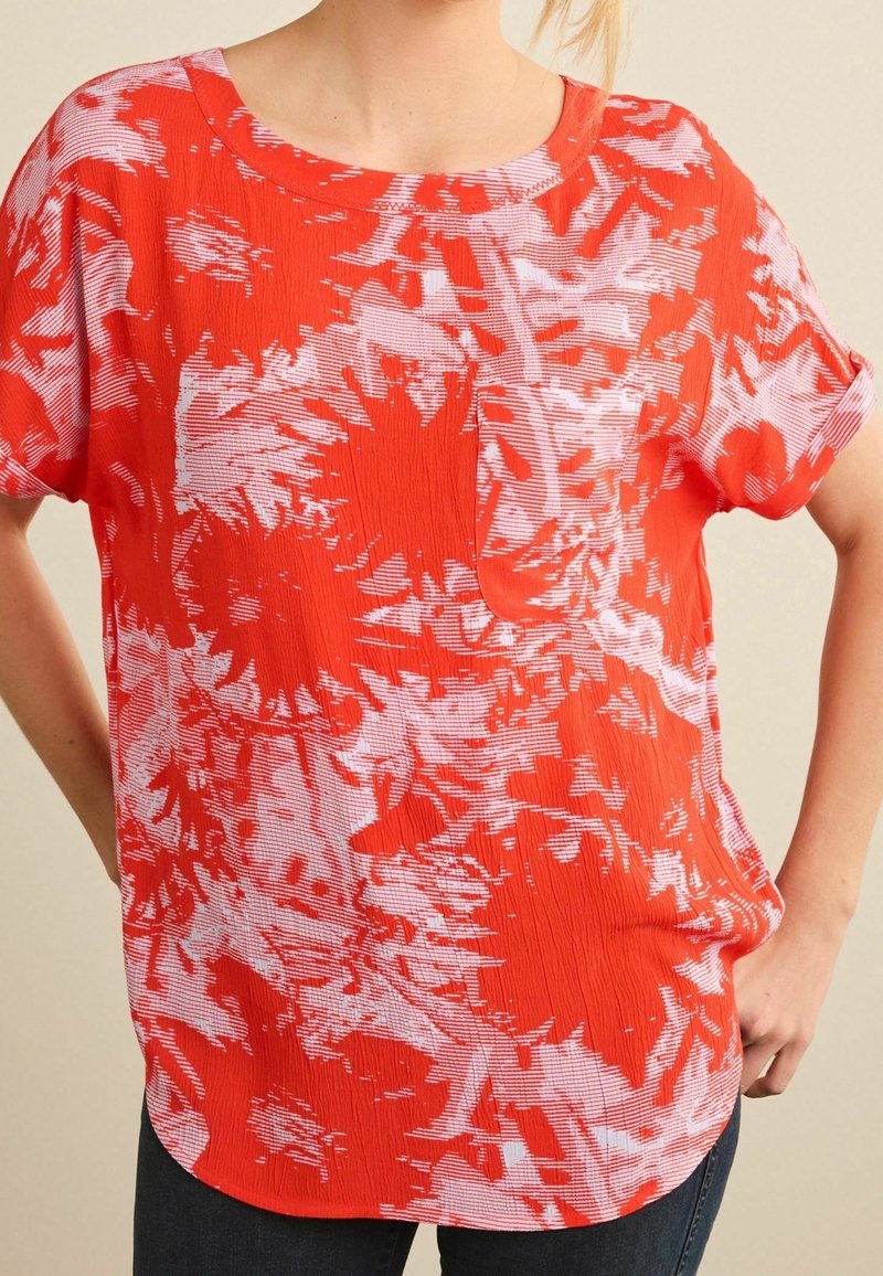 Femme portant un chemisier rouge à manches courtes, ample, avec des motifs floraux abstraits blancs et une petite poche poitrine, associé à un pantalon foncé.