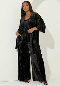 Zwart fluwelen outfit met een wijde broekuitdrukking en losse, vloeiende mouwen. De stof heeft een gepolyesterde, verticale streepstructuur.